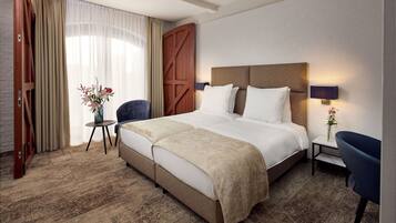 Ein Hotelzimmer mit einem großen Bett, einem kleinen Tisch, einem Sessel, einem Nachttisch mit Lampe und einem großen Fenster mit Vorhängen.
