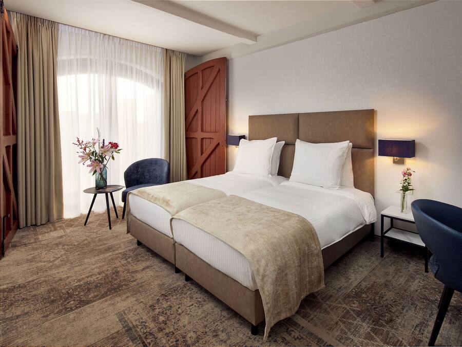Ein Hotelzimmer mit einem großen Bett, einem kleinen Tisch, einem Sessel, einem Nachttisch mit Lampe und einem großen Fenster mit Vorhängen.