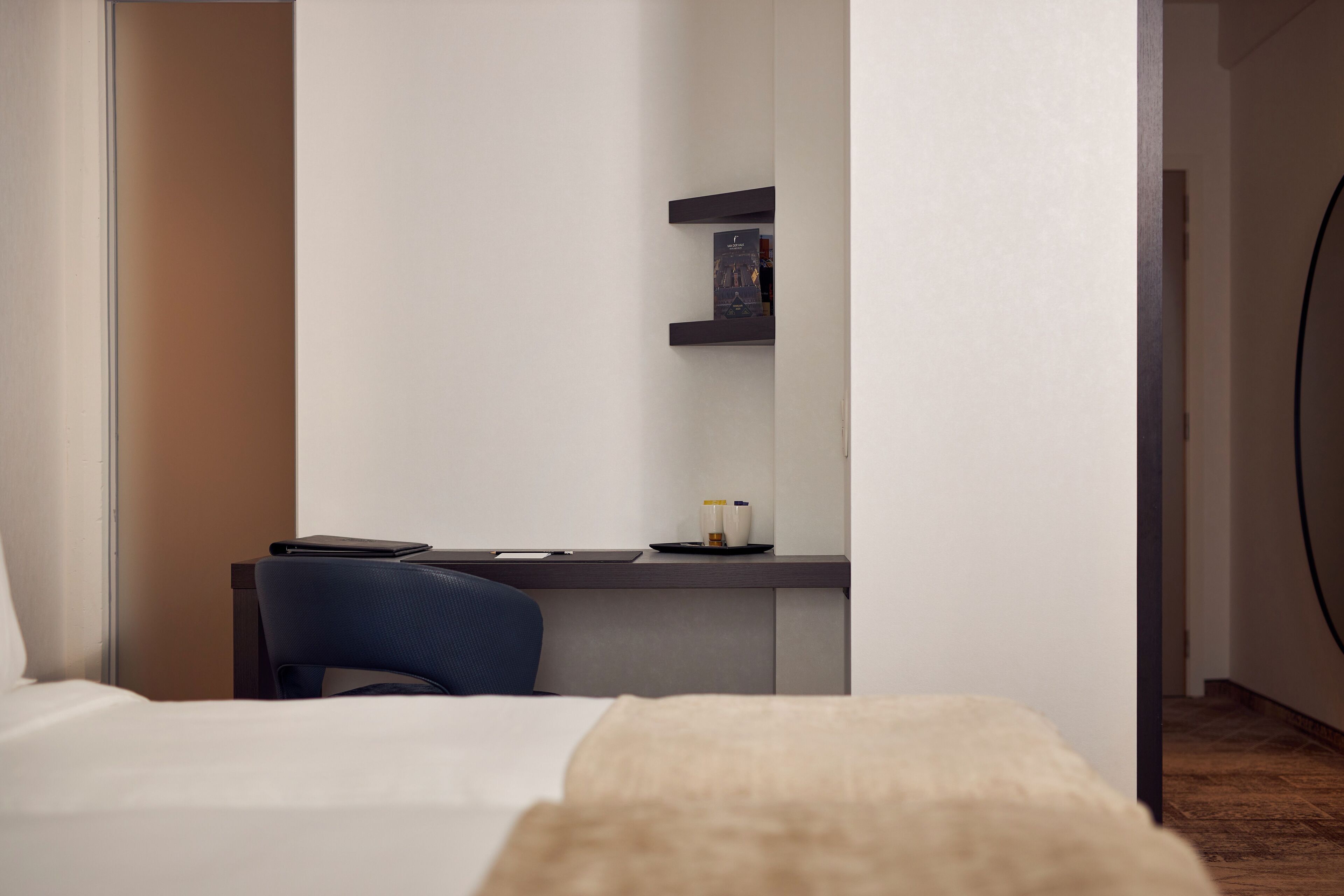 A hotel room with a bed, a desk with a chair, a small shelf with books, and a bedside table with a lamp.