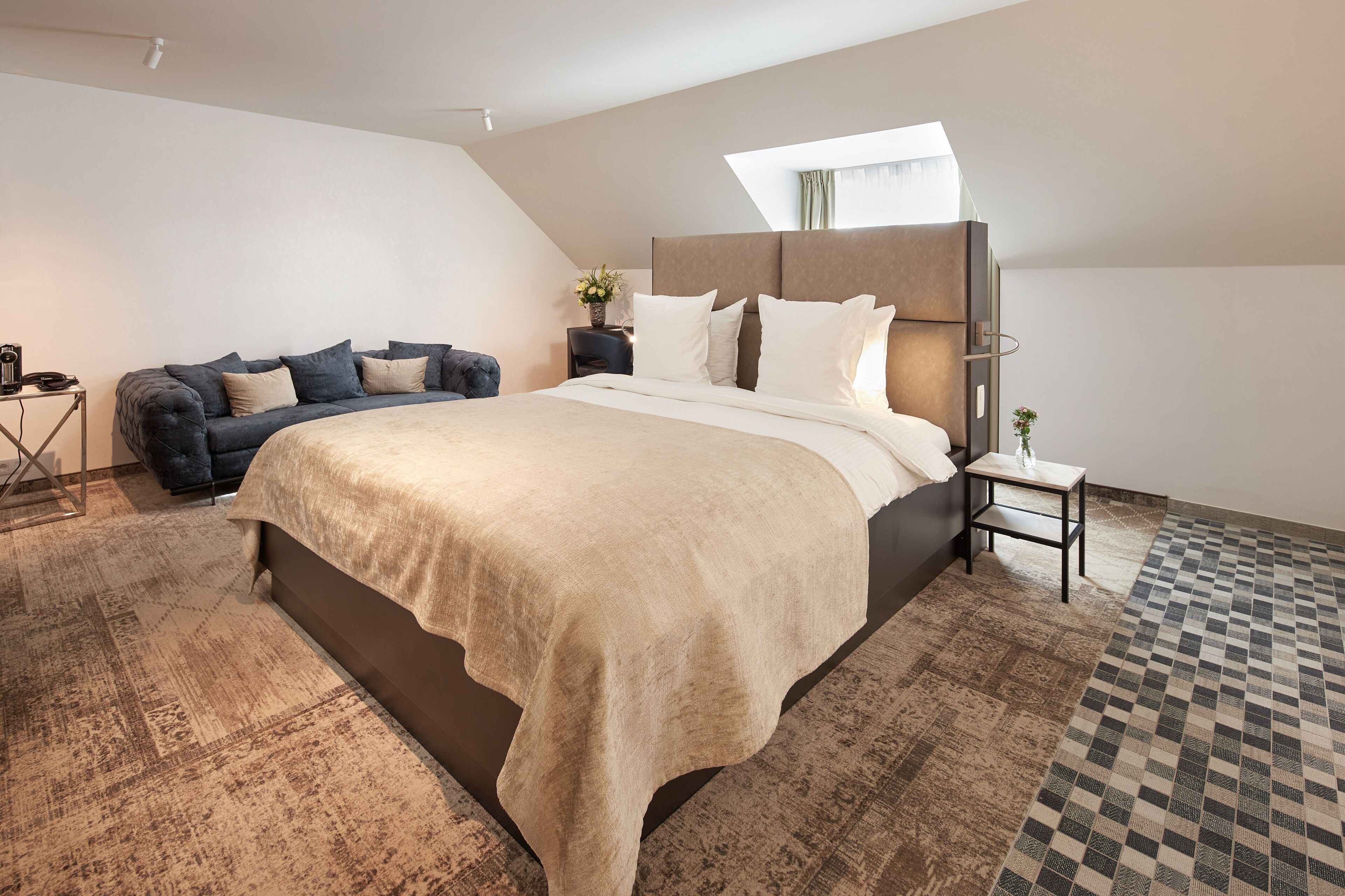 A modern bedroom with a large bed, a dark blue sofa, a small side table, and a patterned rug.