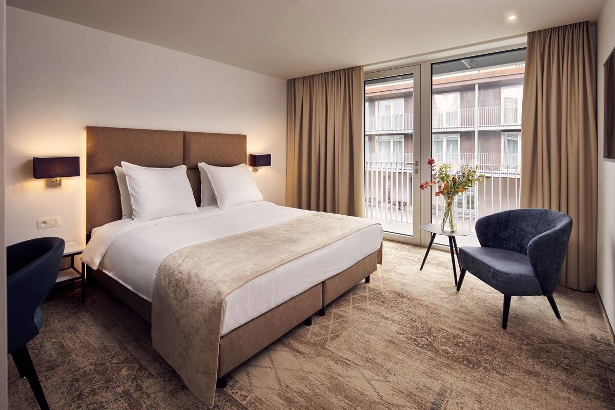 Ein Hotelzimmer mit einem großen Bett, einem Sessel, einem kleinen Tisch mit einer Vase und einem Balkon mit Vorhängen.