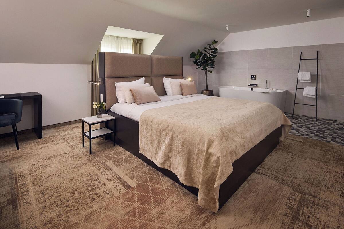 Ein modernes Hotelzimmer mit einem großen Bett, einem Schreibtisch, einem kleinen Beistelltisch, einer Topfpflanze, einer Badewanne und einem Handtuchhalter.