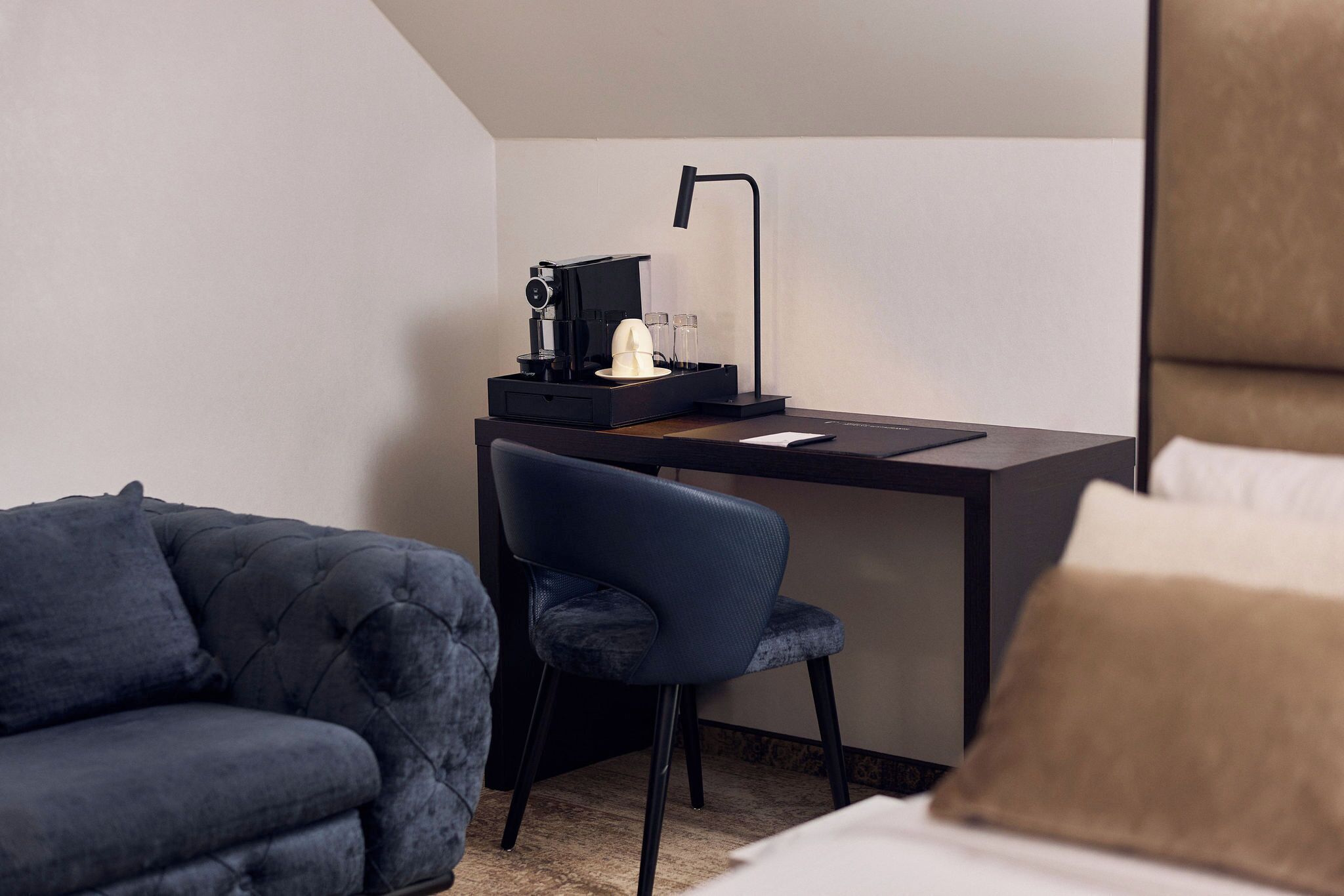 A hotel room with a dark blue upholstered sofa, a matching armchair, a desk with a laptop, a coffee maker, and a lamp.
