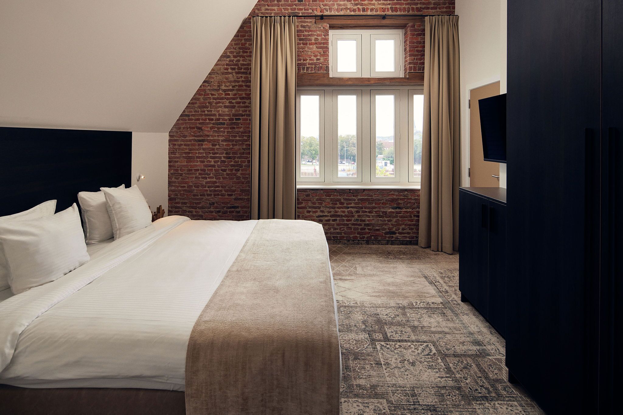 A bedroom with a large bed, a brick wall, a window with curtains, and a TV on a dark cabinet.