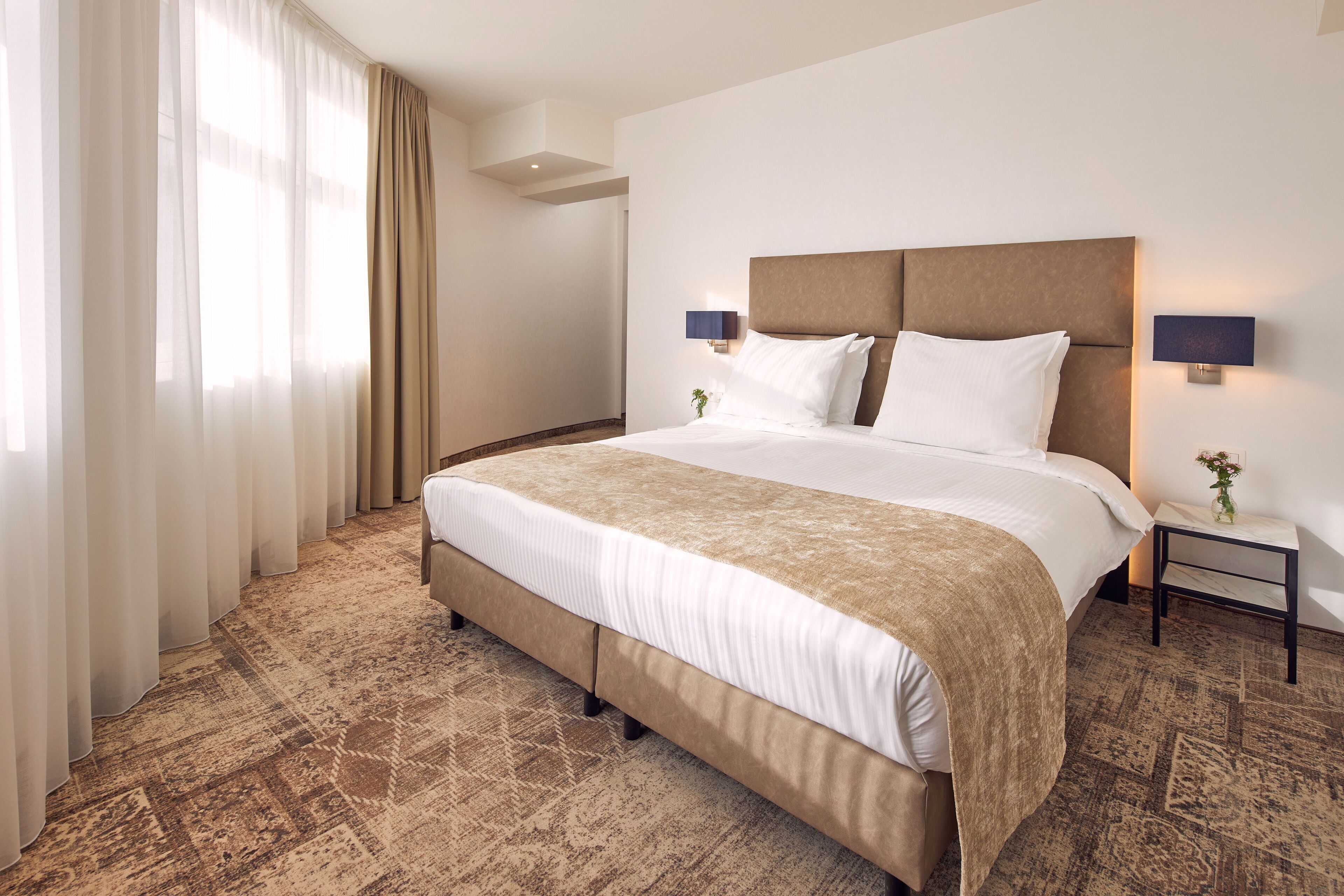 Hotelkamer met een groot bed, een nachtkastje met een lamp, en uitzicht op de omgeving door dunne gordijnen.