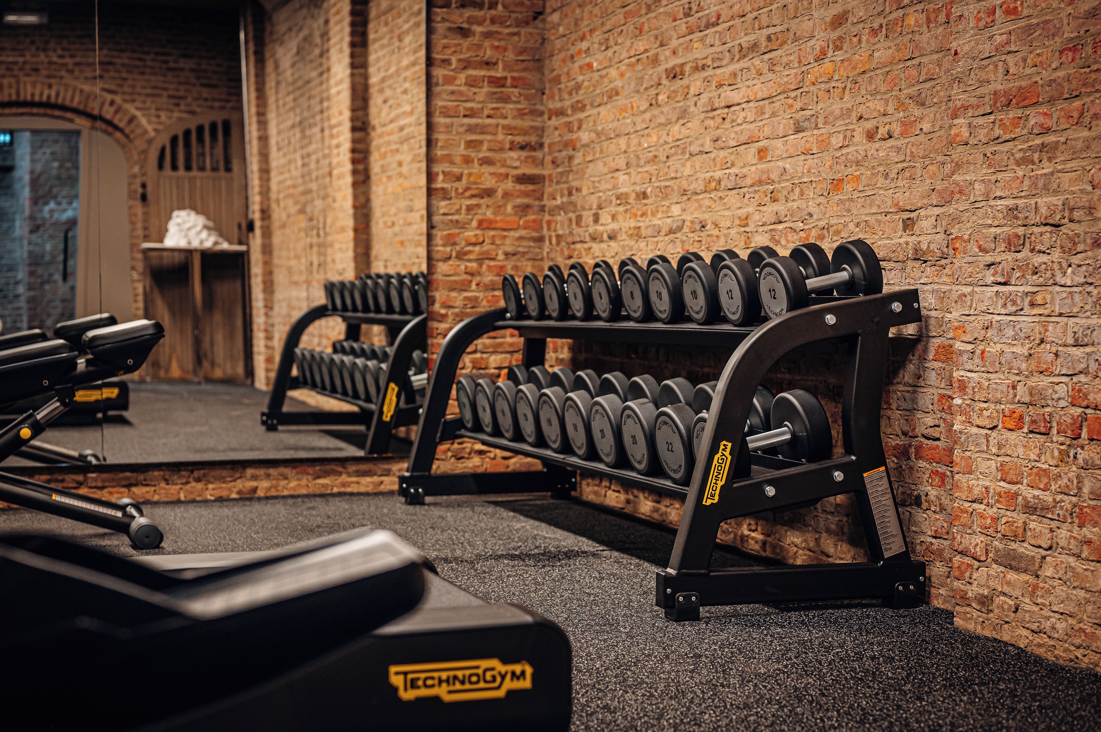 A gym with weightlifting equipment, including dumbbells and benches, against a brick wall.