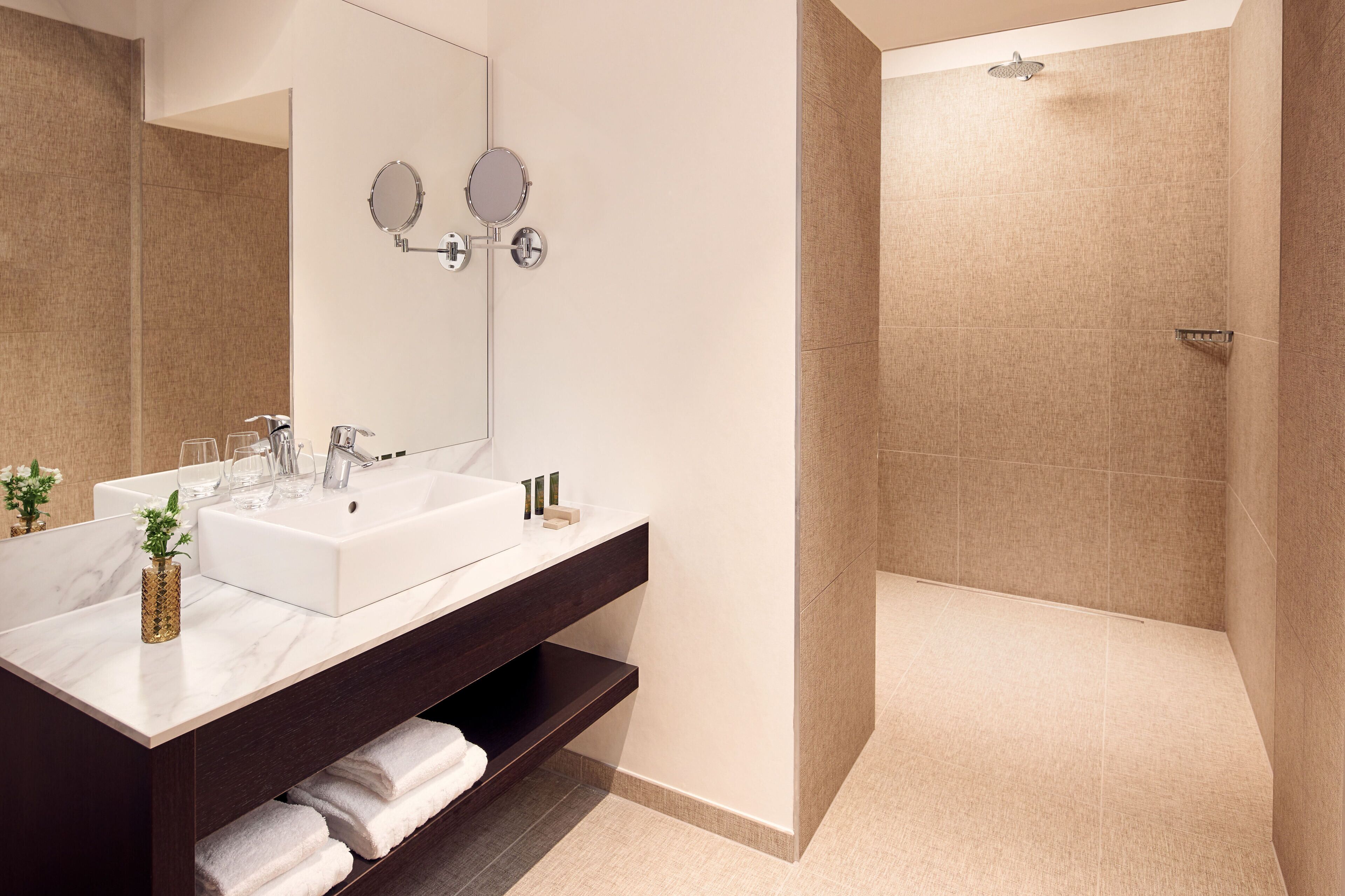 A modern bathroom with a white sink, two mirrors, a shower, and a dark vanity with a towel rack.