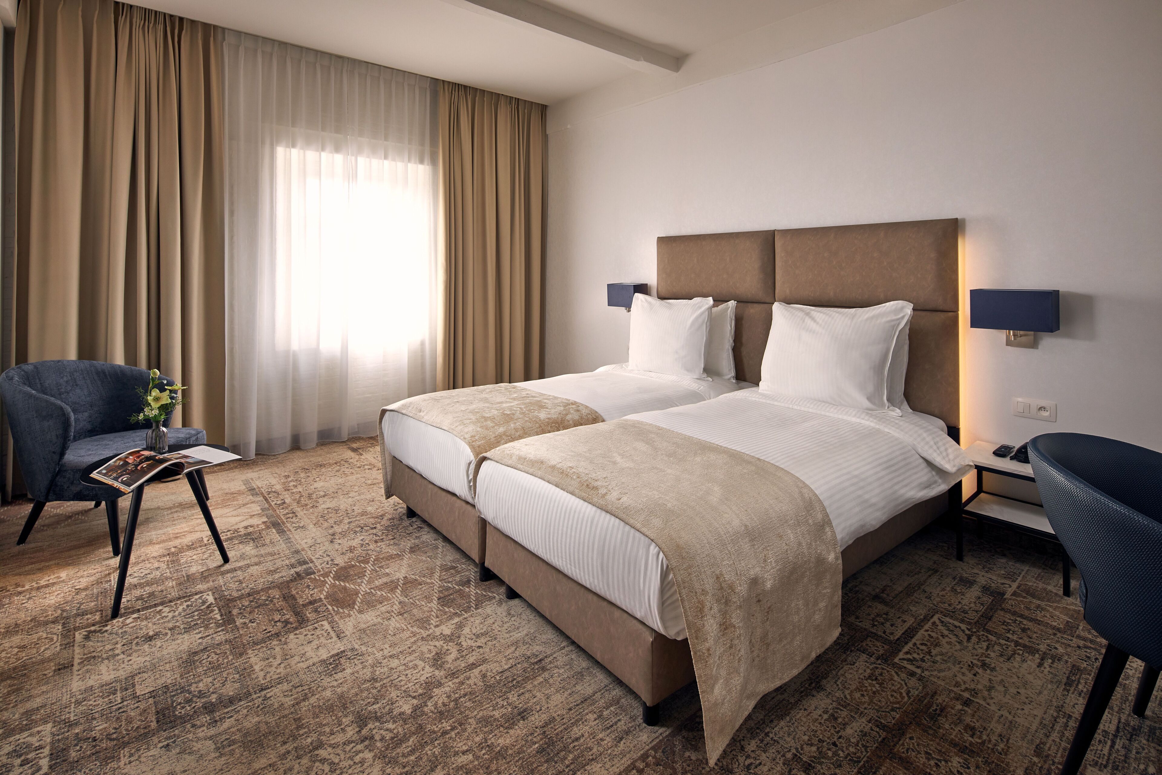 Une chambre d’hôtel avec un lit double, une chaise, une petite table et une grande fenêtre avec des rideaux.