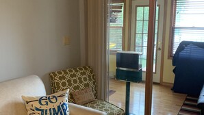 Ein Wohnzimmer mit einem beigen Sofa, einem gemusterten Sessel und einem blauen Sessel. Ein Fenster mit Jalousien und eine zur Außenseite führende Tür sind zu sehen.