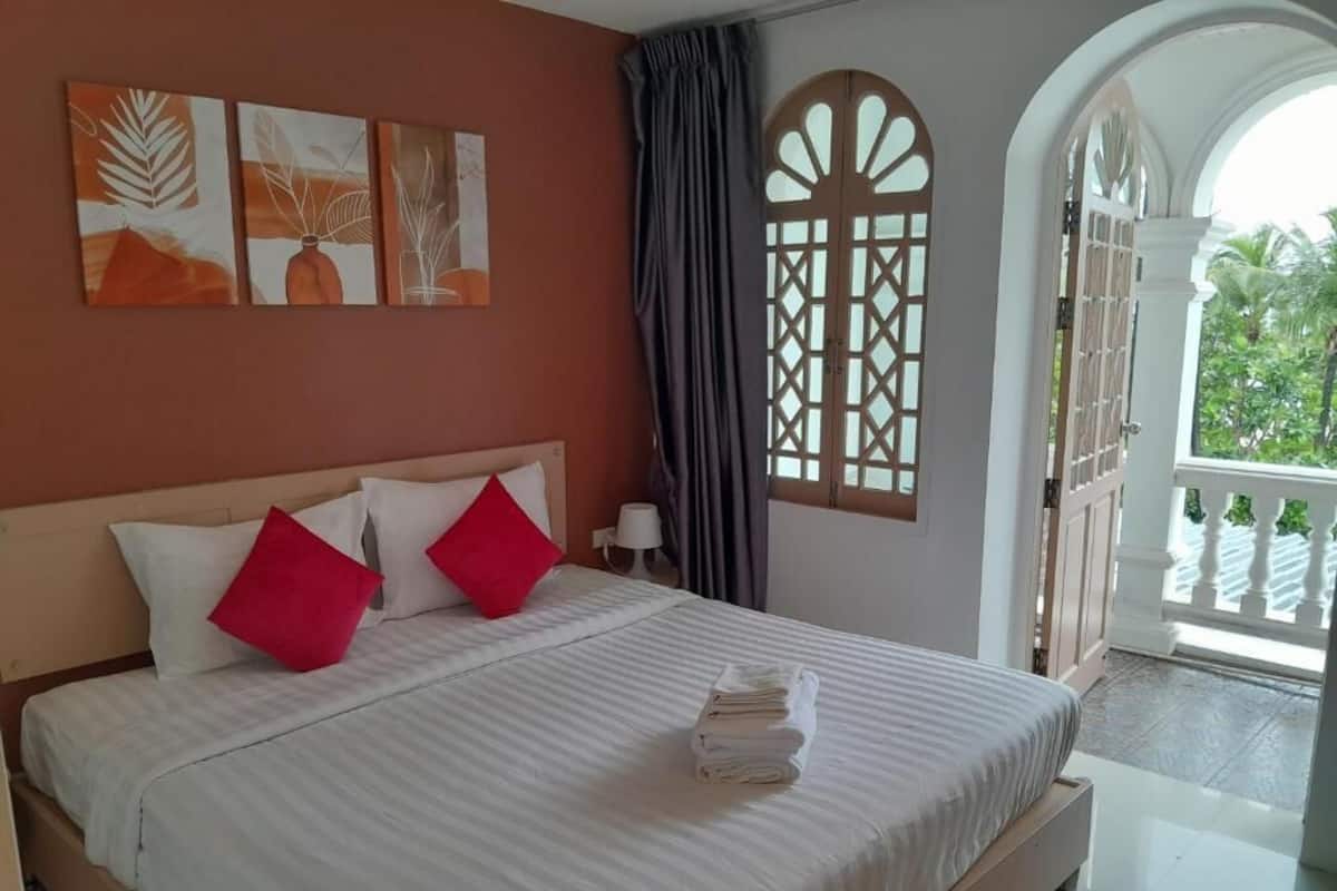 A hotel room with a bed, a view of greenery, a decorative window, and artwork on the wall.