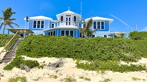 Un edificio grande de varios pisos con una torre destacada, rodeado de vegetación densa y una playa de arena.
