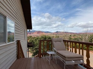 A wooden deck with a cushioned chair and a small table, overlooking a scenic landscape with red rock formations.