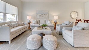 A modern living room with a beige sofa, two armchairs, a coffee table, and a side table with a lamp. There is a painting on the wall and a round mirror on the right.