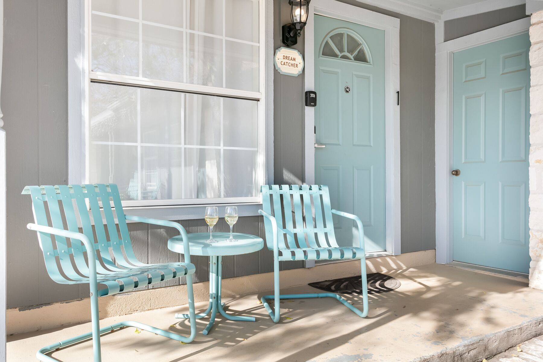 A porch with two light blue chairs and a small table, a light blue door, and a window with white curtains.