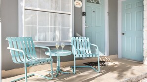 A porch with two light blue chairs and a small table, a light blue door, and a window with white curtains.