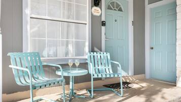 A porch with two light blue chairs and a small table, a light blue door, and a window with white curtains.