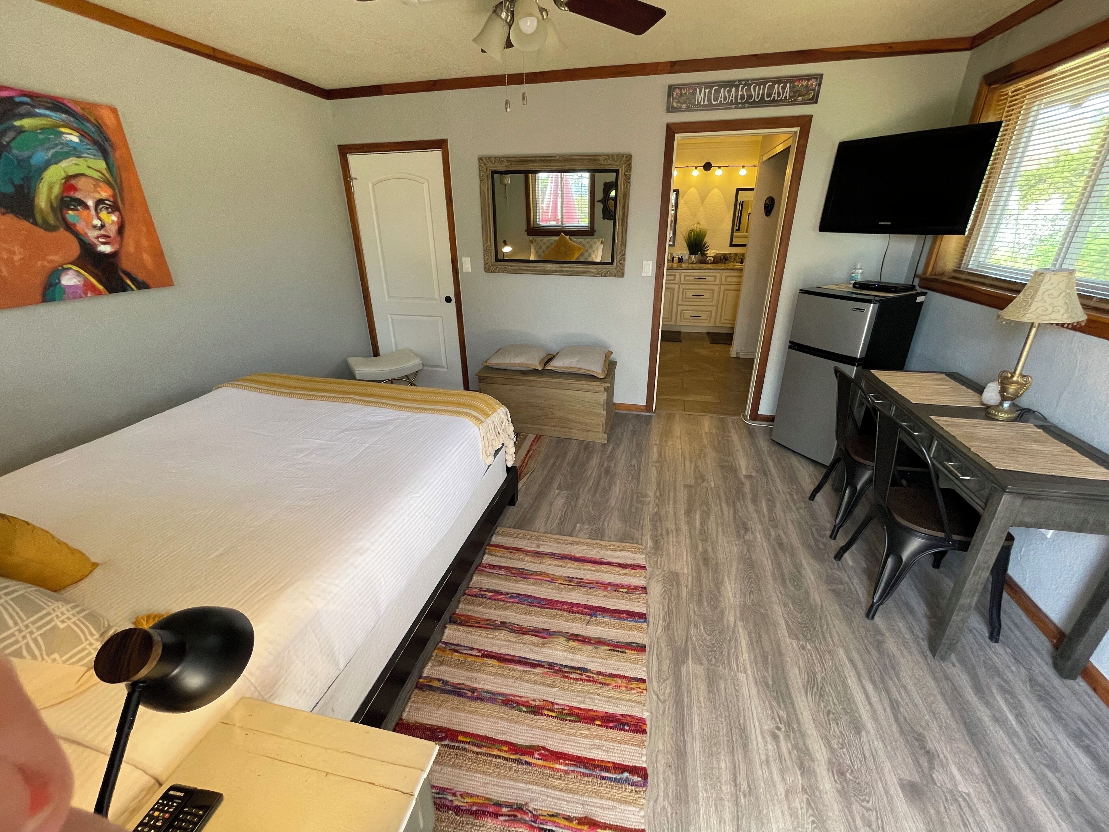 Un dormitorio con cama, escritorio, silla, televisor y un cuadro en la pared.