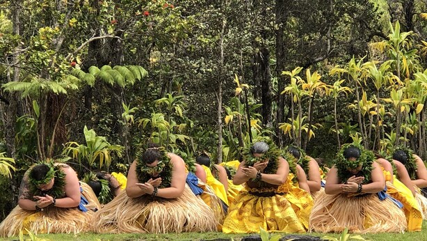 A group of people in traditional attire with headdresses, gathered in a natural setting with dense foliage and a stone wall in the foreground.
