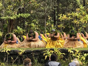 Un groupe de personnes, vêtues de costumes traditionnels et coiffées de couvre-chefs, s'est rassemblé dans un cadre naturel, caractérisé par une végétation luxuriante et un mur de pierre au premier plan.