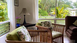 A room with two wicker chairs, a table with a lamp, and a view of greenery outside.