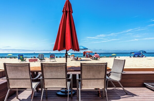 Une terrasse au bord de la plage, avec des chaises en osier et un parasol rouge, donnant sur une plage de sable où se trouvent des personnes et d’autres parasols.