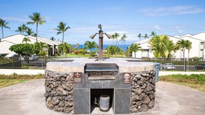 A barbecue grill with a stone base, surrounded by a fence, palm trees, and a view of the ocean.
