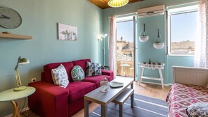A cozy room with a red sofa, a small table, a hanging plant, and a view of buildings through a window. - St Spyridon Cosy Apartment By Konnect (Corfu)