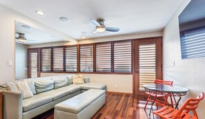 A living room with a sectional sofa, a glass-top dining table with red chairs, wooden blinds on the windows, and recessed ceiling lights. - Aloha Condos, Napili Bay Resort, Condo 105, Beachfront (Lahaina)