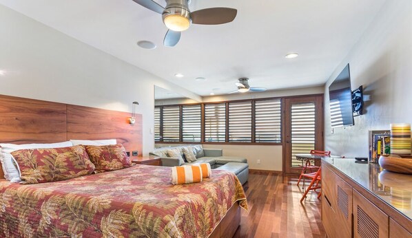 Une chambre moderne avec un grand lit, un bureau et une chaise. Elle est dotée de fenêtres avec stores, d’un ventilateur de plafond et d’un téléviseur fixé au mur.