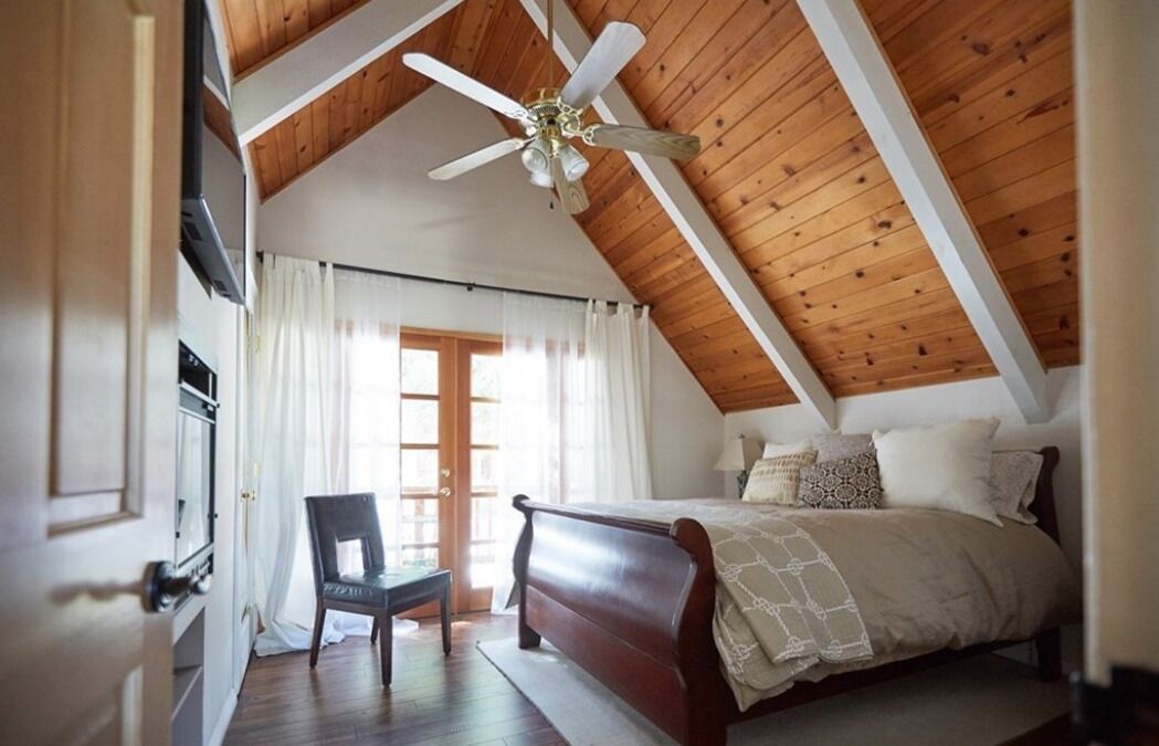 Une chambre avec un plafond en bois, un lit, une chaise et une fenêtre avec des rideaux.