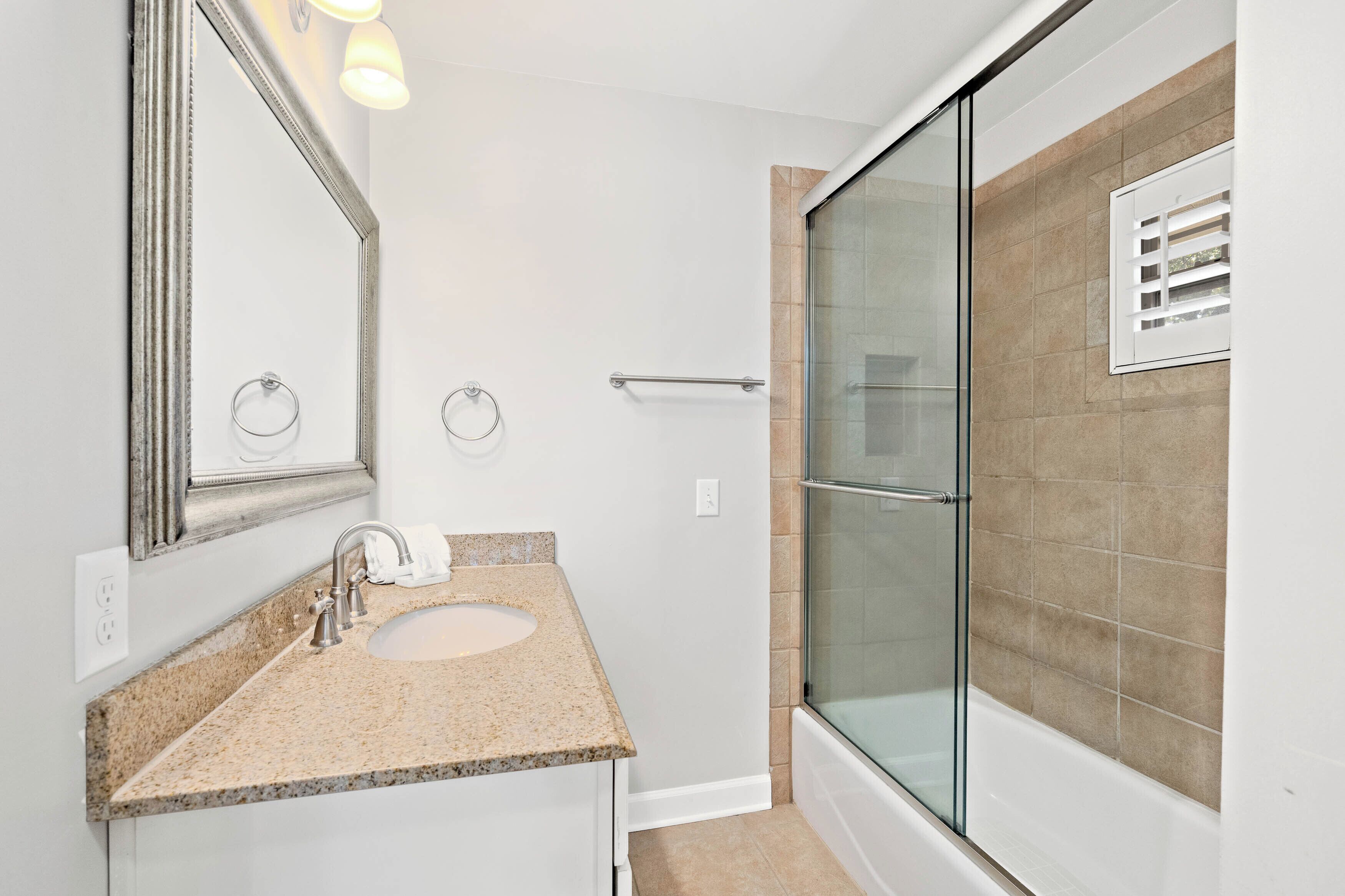 A bathroom with a granite countertop sink, a large mirror, a glass shower enclosure, and beige tiles.