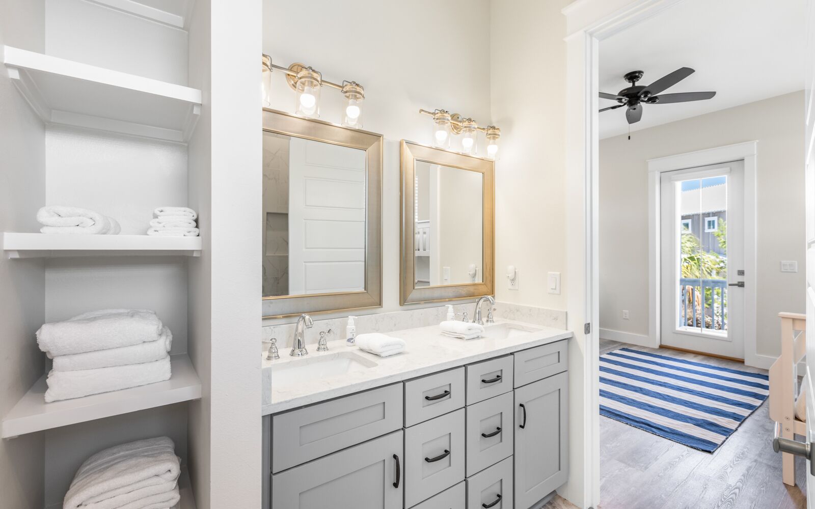 A bathroom with two sinks, a large mirror, and a ceiling fan. There are shelves with folded towels and a blue and white striped rug on the floor.