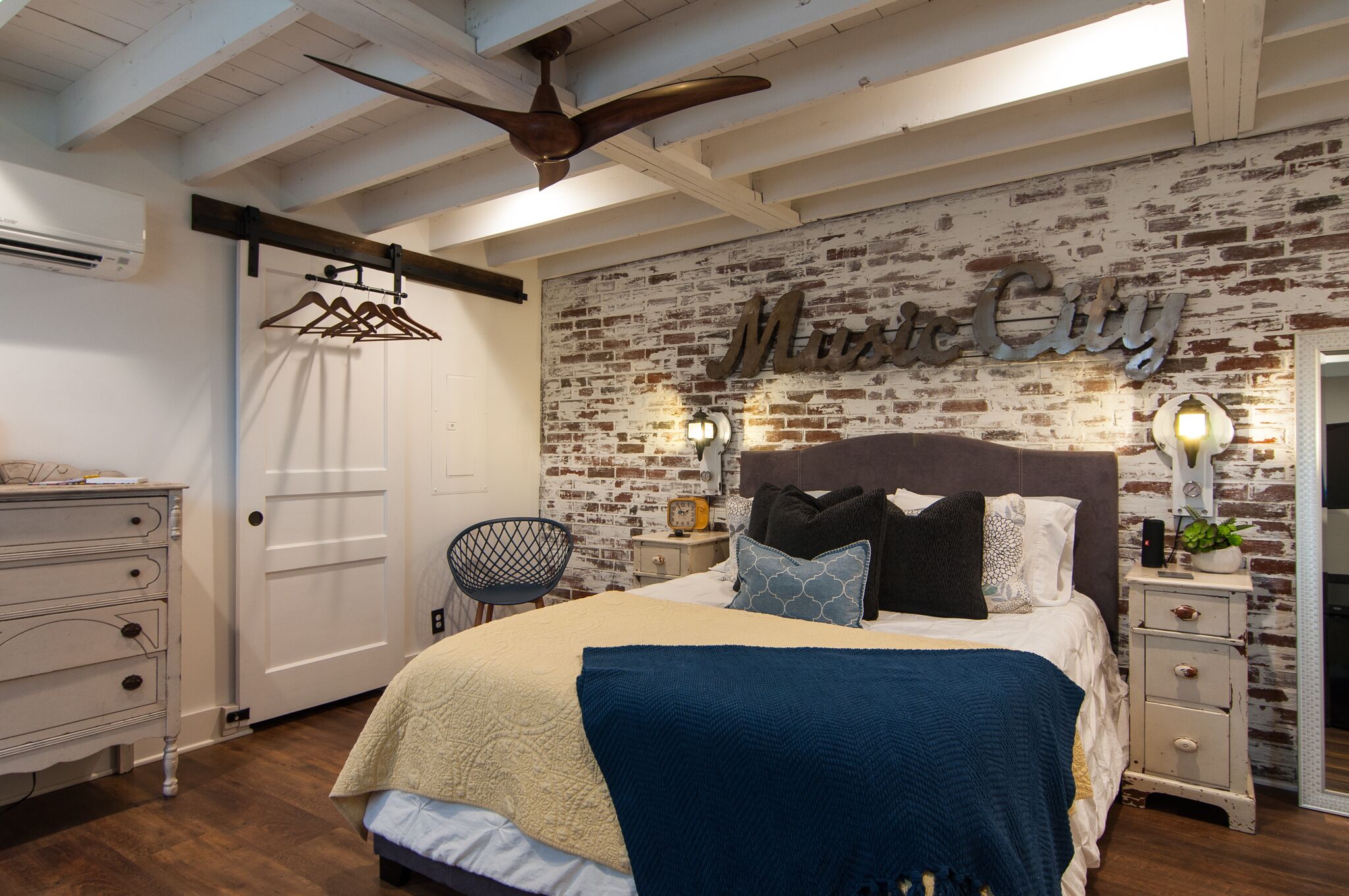 A bedroom with a brick wall, a bed with multiple pillows, a nightstand with a lamp, and a ceiling fan.
