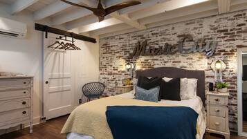 A bedroom with a brick wall, a bed with multiple pillows, a nightstand with a lamp, and a ceiling fan.