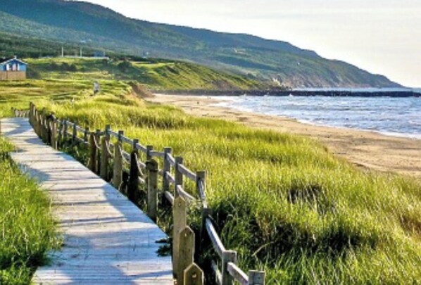 A wooden boardwalk leading to a beach with grassy areas and a hill in the background. - House on the lake. Large home with cathedral ceiling, (Inverness, Cape Breton)