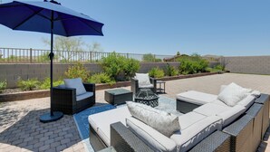 An outdoor patio with wicker furniture, a blue umbrella, and a brick wall in the background. - Group-Friendly Estrella Gem: Hike, Cheer & Explore (Goodyear)