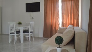 A living room with a beige sofa, a white dining table with two chairs, a potted plant, and a flat-screen TV mounted on the wall. - PERAK apartments Tivat (Tivat)