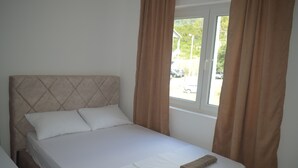 A neatly made bed with white linens and two brown towels, a window with curtains, and a view of a parking area outside. - PERAK apartments Tivat (Tivat)