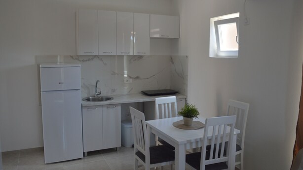 A compact kitchen with white cabinets, a refrigerator, a sink, and a dining table with chairs. - PERAK apartments Tivat (Tivat)