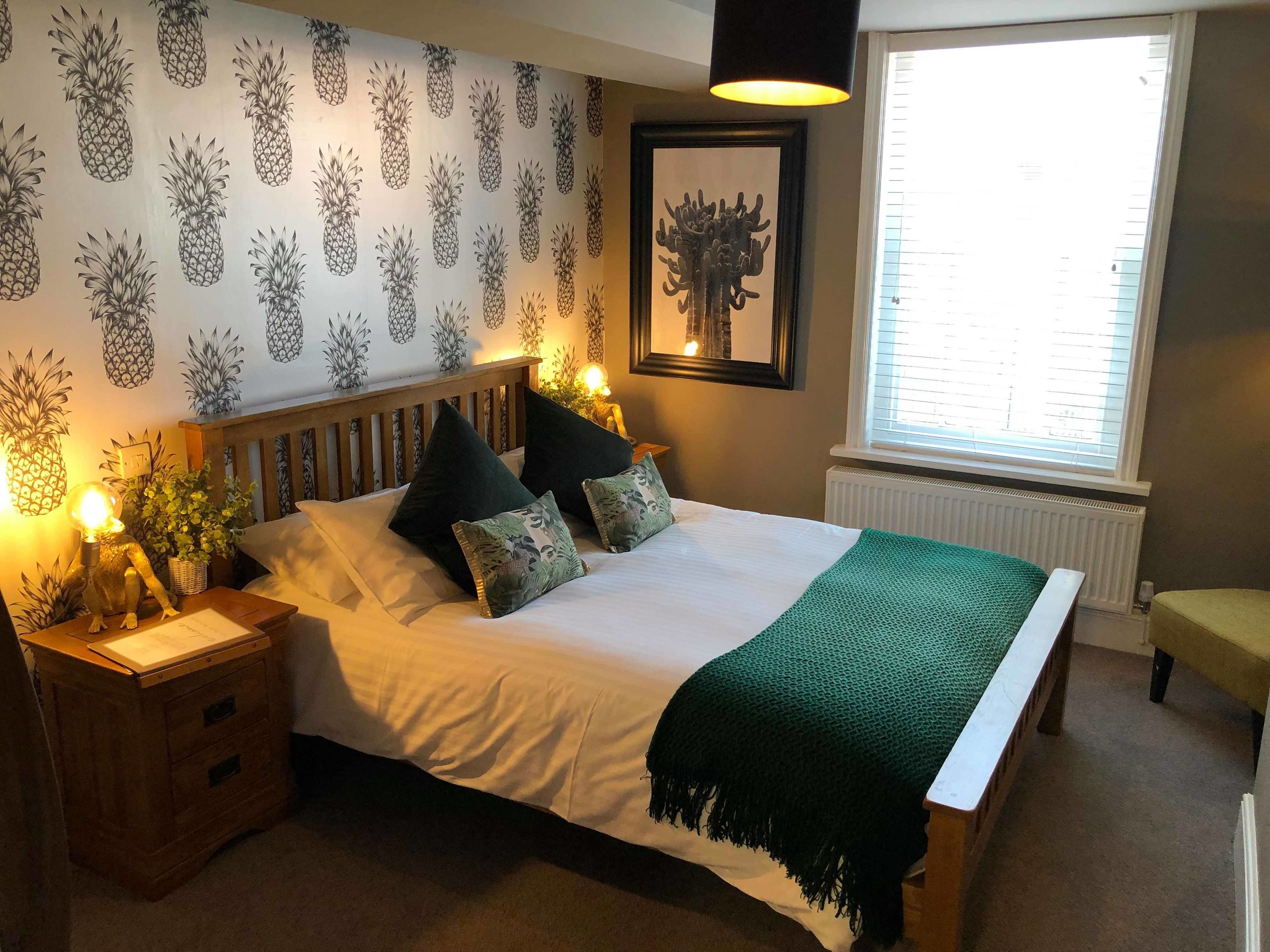Ein Schlafzimmer mit einem Bett, Kissen, einem Nachttisch mit Lampe, einer grünen Decke, einer Wand mit Ananas-Mustern und einem Fenster mit Jalousien.