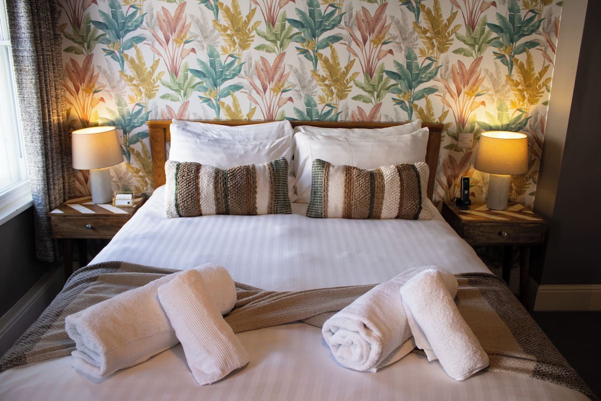 A neatly made bed with white linens, two pillows, and two rolled towels.