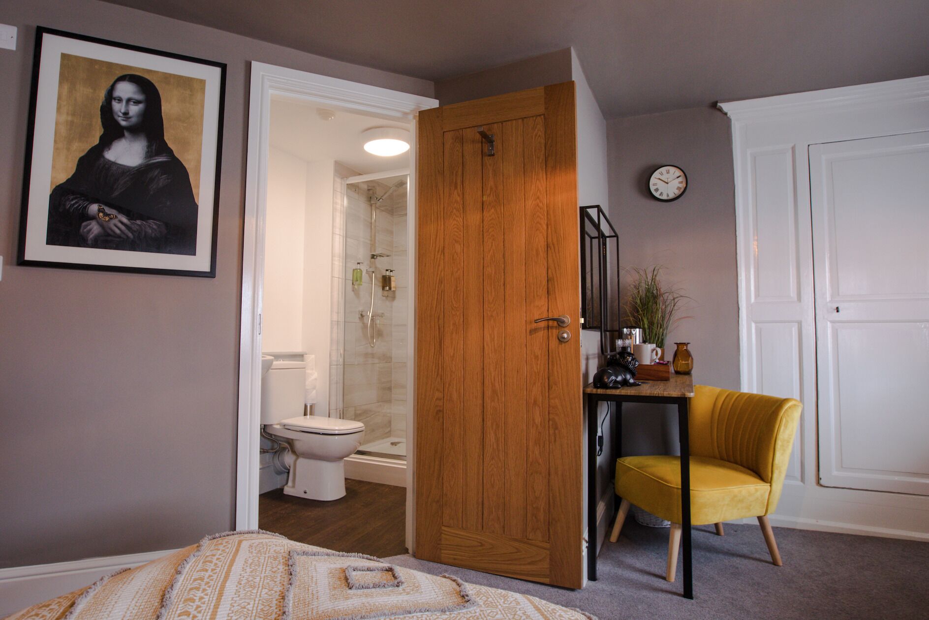 Ein Schlafzimmer mit einem Bett, einem Holzschrank, einem gelben Sessel, einem Badezimmer mit Toilette und einem gerahmten Kunstwerk an der Wand.