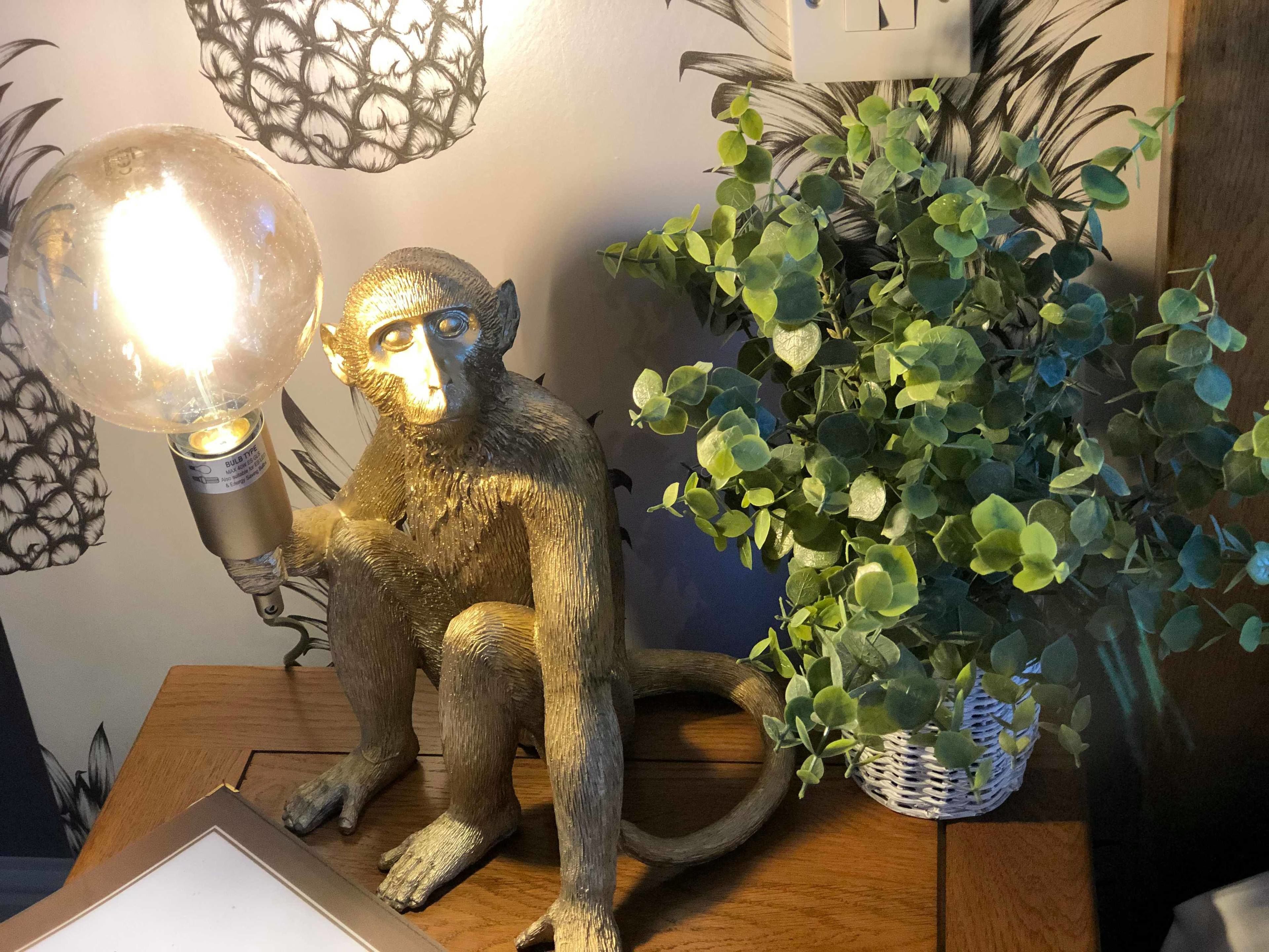 A monkey sculpture with a light fixture on its back, a potted plant, and a wall with a leaf pattern.