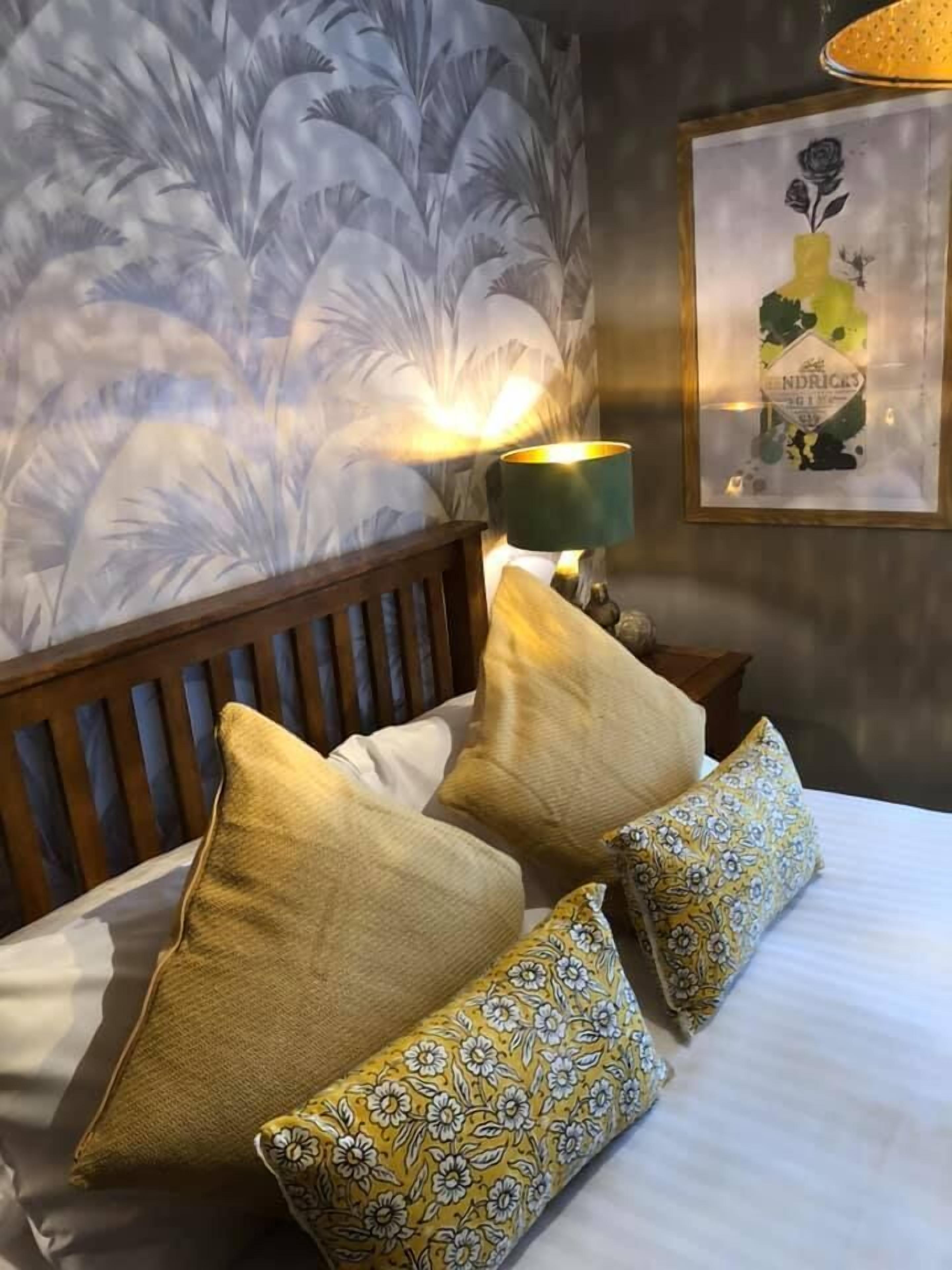 Un lit bien fait, agrémenté d’oreillers à motifs floraux, d’une tête de lit en bois et d’un mur recouvert d’un grand papier peint à fleurs.