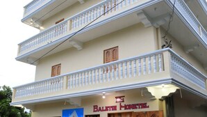 Um edifício de três andares com varandas, uma placa com a inscrição "Balefish Honavel" e uma pequena área de entrada com uma porta e um banco.