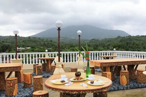 Un área de comedor al aire libre con mesas redondas y taburetes, rodeada por un balcón de balaustradas blancas, frente a un fondo de vegetación exuberante y una montaña.