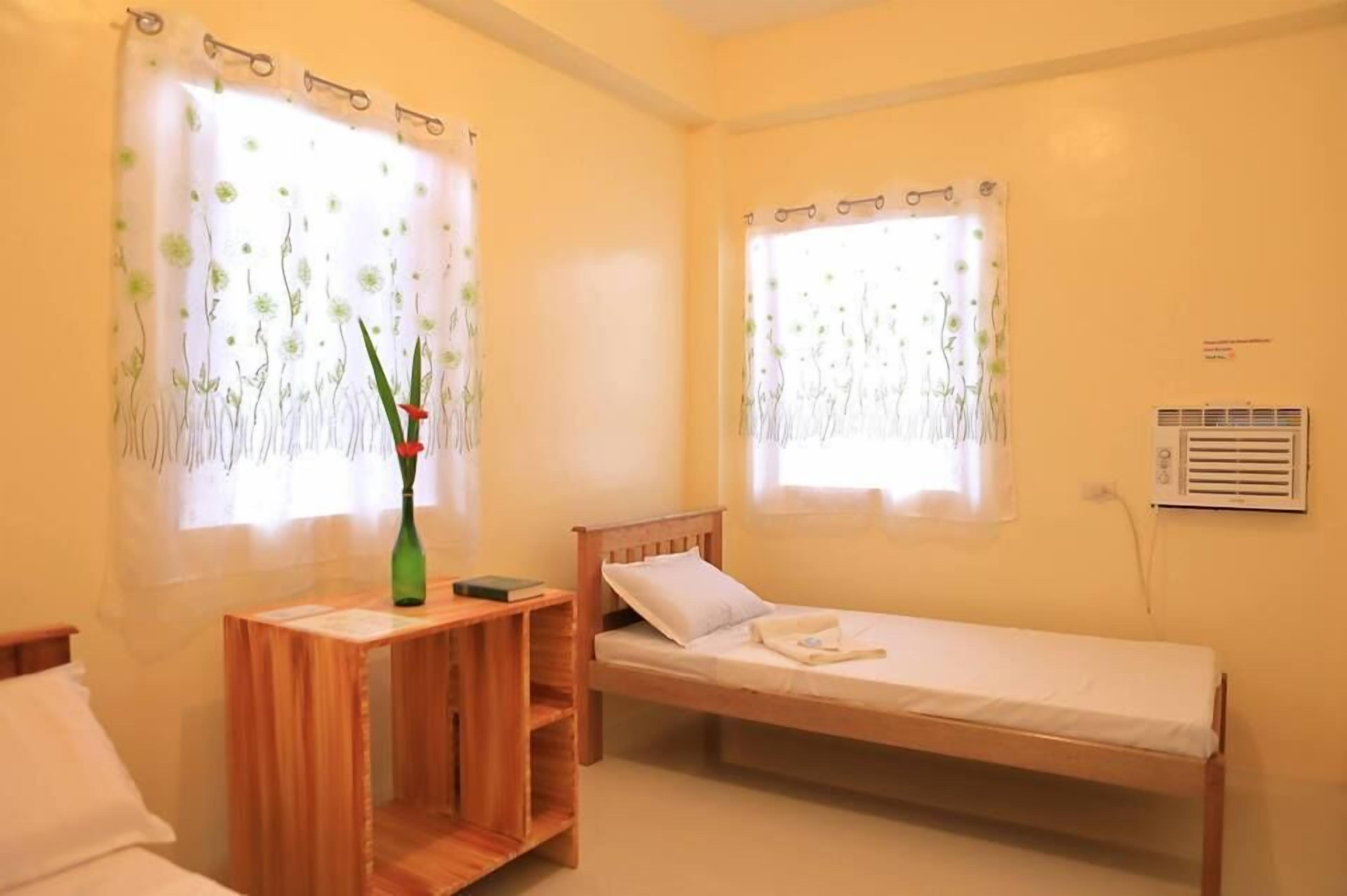 A small, clean room with two single beds, a wooden nightstand, a green vase, and a window with a floral curtain.