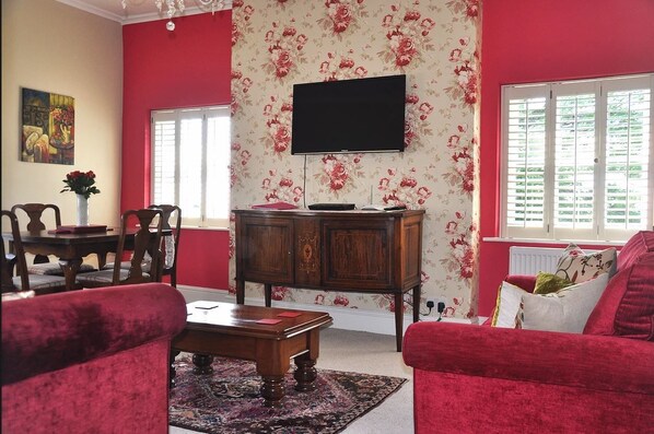 A living room with a red sofa, a wooden coffee table, a floral wallpaper, a flat-screen TV, and a dining area with a red table and chairs.
