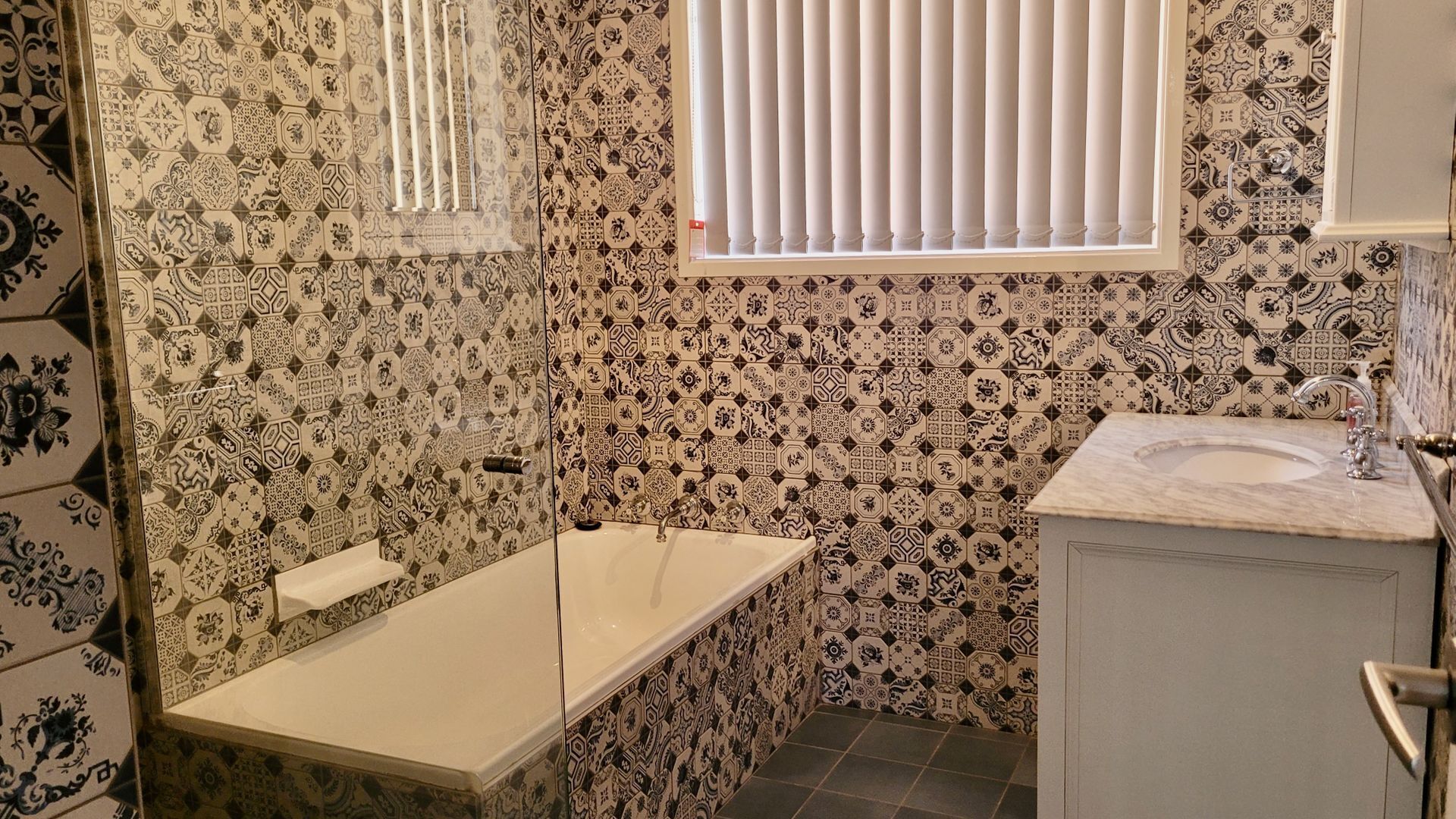 Une salle de bain avec des carreaux décorés, une baignoire, un lavabo et une fenêtre avec des stores.