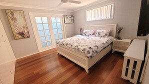 Un dormitorio con suelo de madera, cama con ropa de cama floral, mesita de noche, ventilador de techo y ventana con persianas.